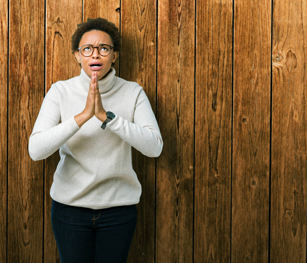 Νέοι όμορφη αφρικανική αμερικανική γυναίκα που φοράει γυαλιά, πέρα από το απομονωμένο υπόβαθρο επαιτεία και προσεύχεται με τα χέρια μαζί με ελπίδα έκφραση προσώπου πολύ συναισθηματική και ανησυχούν. Ζητώντας συγχώρεση. Έννοια της θρησκείας. - Φωτογραφία, εικόνα