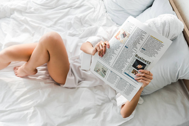 top view of woman with towel on head and obscure face lying in white bed and reading business newspaper - Fotoğraf, Görsel