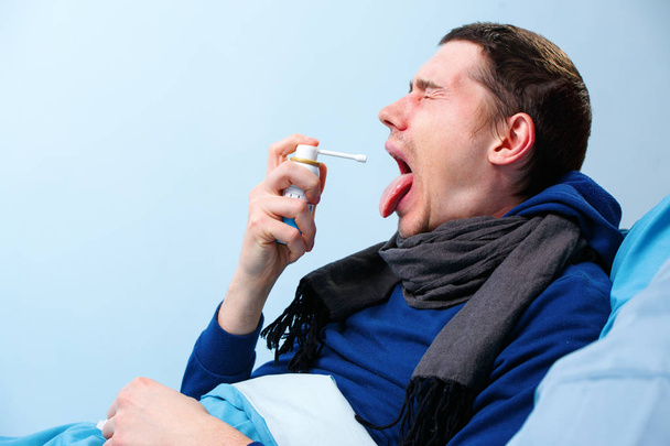 Φωτογραφία από την πλευρά του άρρωστου άντρα με κασκόλ χρησιμοποιώντας σπρέι με φάρμακο για το λαιμό που βρίσκεται στο κρεβάτι - Φωτογραφία, εικόνα