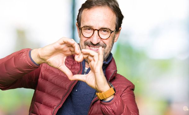 Hombre guapo de mediana edad con gafas y abrigo de invierno sonriendo en el amor mostrando el símbolo del corazón y la forma con las manos. Concepto romántico
. - Foto, imagen