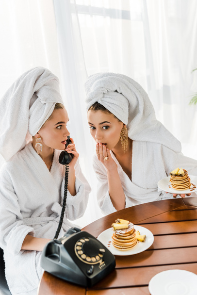 stylish shocked women in bathrobes and jewelry with towels on heads talking on retro telephone while having breakfast - 写真・画像