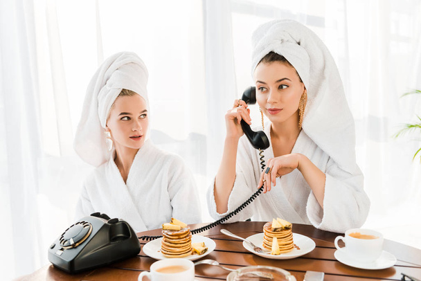 стильные женщины в халатах и украшениях с полотенцами на головах с помощью ретро-телефона во время завтрака с блинами
  - Фото, изображение