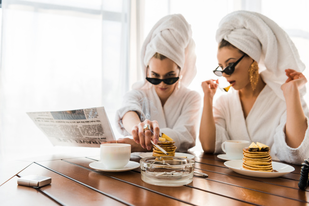 selective focus of stylish women in bathrobes, sunglasses and jewelry with towels on heads smoking cigarette and reading newspaper while eating pancakes - Photo, Image
