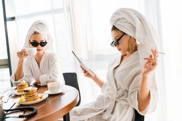 stylish women in bathrobes, sunglasses and jewelry with towels on heads smoking cigarette and reading newspaper at morning - Foto, Bild