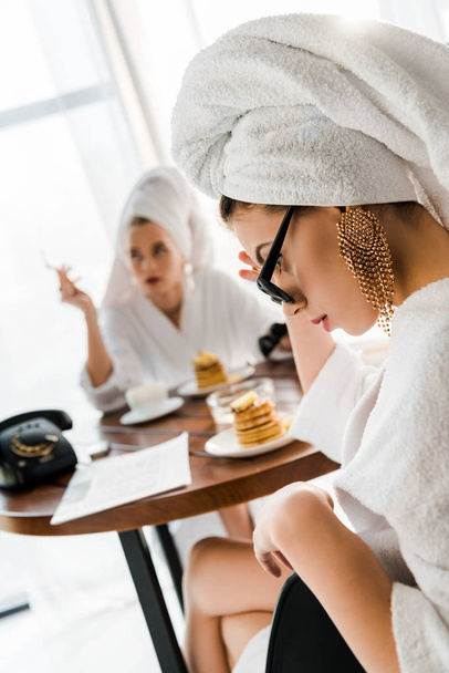 selektiver Fokus der aufgebrachten stilvollen Frau in Bademantel, Sonnenbrille und Schmuck mit Handtuch auf dem Kopf am Tisch sitzend - Foto, Bild