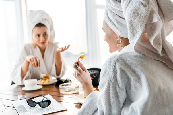 selektywne skupienie uśmiechniętej stylowej kobiety w szlafroku i biżuterii z ręcznikiem na głowie siedzi przy stole i rozmawia z przyjacielem - Zdjęcie, obraz