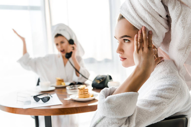 selective focus of exhausted stylish woman in bathrobe and jewelry with towel on head touching head while emotional friend talking on phone - Фото, изображение