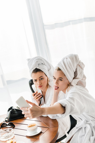 selective focus of stylish women in bathrobes and jewelry with towels on heads talking on retro phone and grimacing while taking selfie on smartphone - Photo, Image