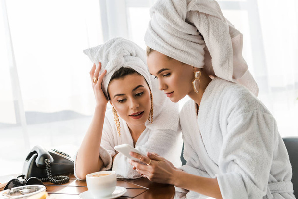 stylish women in bathrobes and jewelry with towels on heads using smartphone - Фото, изображение