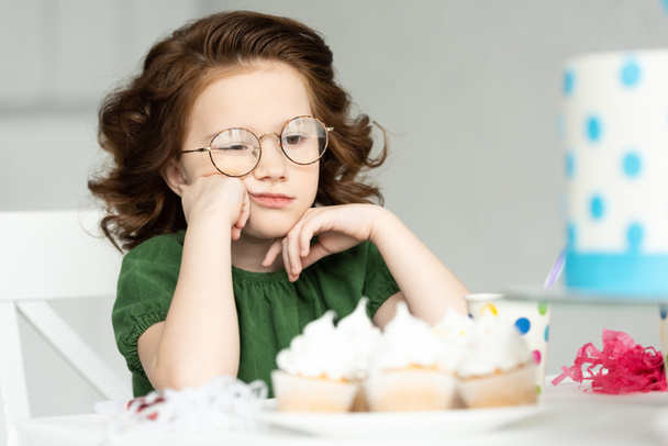 ihana kyllästynyt lapsi pönkittää leuka istuessaan pöydässä cupcakes
 - Valokuva, kuva
