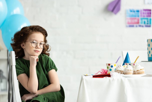 sad kid sitting at table with cupcakes and propping chin during birthday celebration - Foto, Imagen