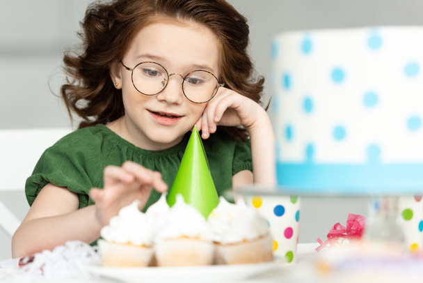 entzückende Preteen sitzt am Tisch und schaut sich Cupcakes während der Geburtstagsfeier zu Hause an - Foto, Bild