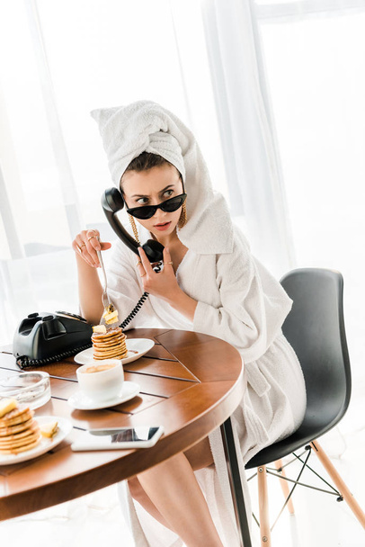 stylish woman in bathrobe, sunglasses and jewelry with towel on head talking on retro telephone while eating pancakes - Fotoğraf, Görsel