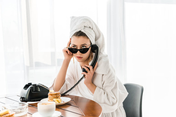 mujer elegante en albornoz, gafas de sol y joyas con toalla en la cabeza hablando por teléfono retro durante el desayuno
 - Foto, Imagen