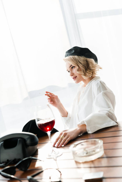 elegáns álmodozó szőke nő, fekete Beret és fehér inget dohányzás cigaretta közelében üveg vörösbor és retro telefon - Fotó, kép