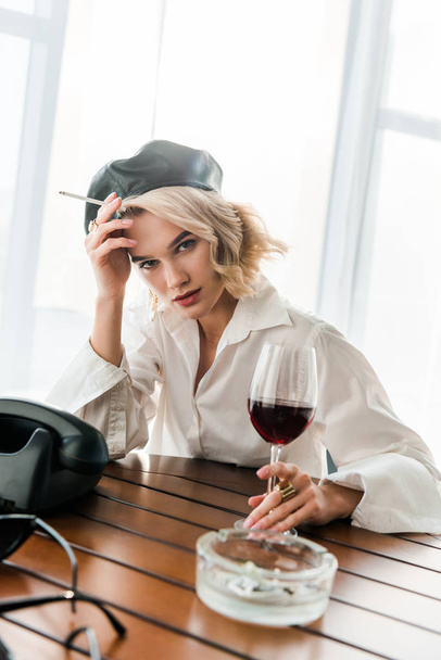 elegant blonde woman in black beret and white shirt  holding cigarette and glass with red wine  - Foto, afbeelding