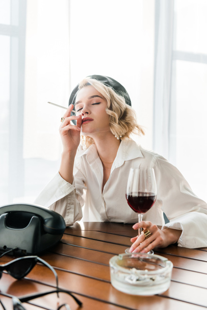elegantní zasněný blondýnka v černém baretu a bílé košili s cigaretou a sklenkou s červeným vínem  - Fotografie, Obrázek