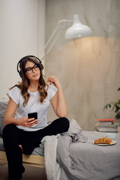 Attractive Caucasian brunette sitting on the bed in pajamas with headphones on head and smart phone in hand. On bed breakfast. Bedroom interior. - 写真・画像