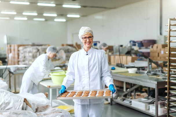 Uśmiechnięta blond kaukaski pracownik w sterylnym mundurze i okularach stojących i trzymając tacę z ciasteczkami. Wnętrze fabryki żywności. W tle inni pracownicy pracujący. - Zdjęcie, obraz
