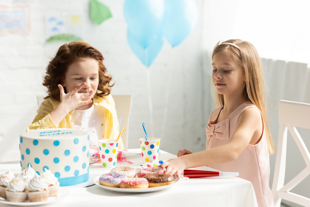 rozkošné děti sedící na stranovém stole s dortíky a koláčem na narozeninové oslavě - Fotografie, Obrázek
