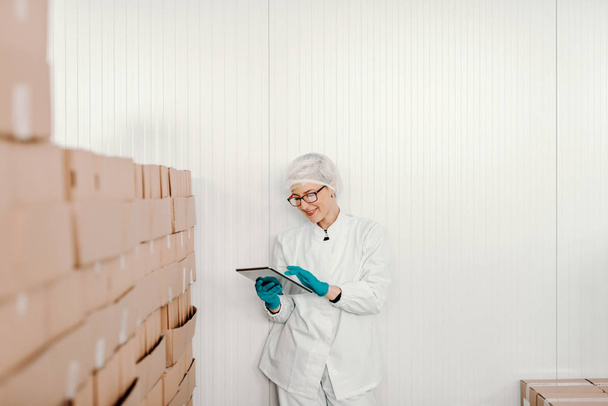 Blondynka pracownica w sterylnych uniform za pomocą tabletu logistyczne chwilę stojąc obok pola w fabryce żywności. - Zdjęcie, obraz