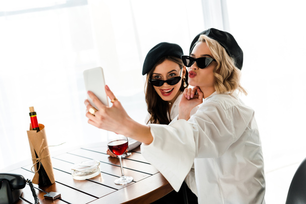 κομψή μελαχρινή και ξανθιά γυναίκες σε μαύρους μπερέδες και γυαλιά ηλίου πίνοντας κόκκινο κρασί και παίρνοντας selfie, ενώ μουτρώνουν τα χείλη και να προεξέχουν γλώσσα  - Φωτογραφία, εικόνα