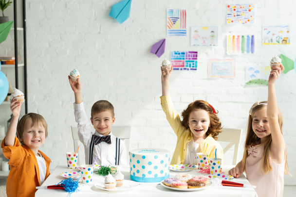 entzückende Kinder sitzen am Tisch und halten Cupcakes, während sie zusammen Geburtstag feiern - Foto, Bild