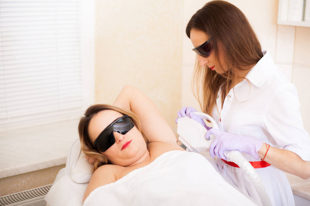 Body Care. Beautiful girl lies in the office of a beautician in goggles from laser irradiation, the hands of the doctor remove hair on hands with a laser device - Photo, Image