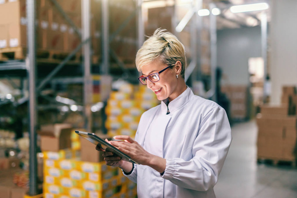 Young Caucasian blonde woman in white uniform using tablet in warehouse. - Photo, Image