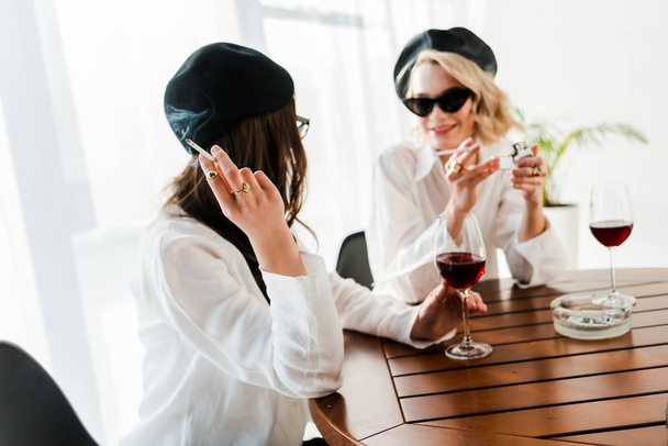 黒いベレーとサングラスを着たブルネットとブロンドの女性が赤ワインを飲み、話し、タバコを吸う - 写真・画像