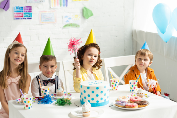 діти у вечірніх капелюхах сидять за столом і дивляться на камеру під час святкування дня народження
 - Фото, зображення