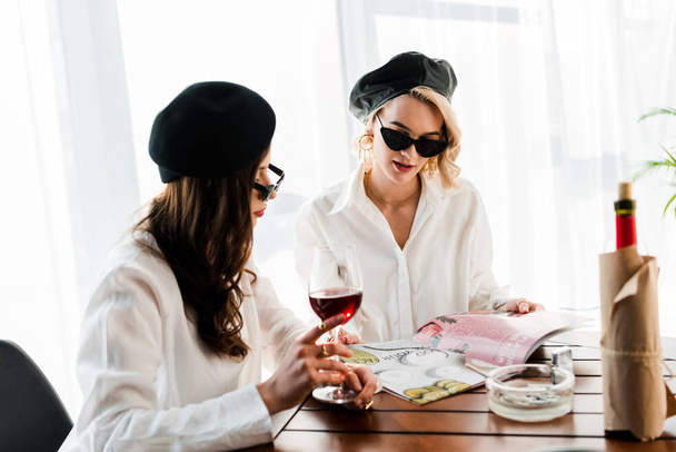 brunette and blonde women in black berets and sunglasses drinking red wine and reading magazine  - Zdjęcie, obraz