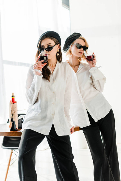 elegant brunette and blonde friends in black berets and sunglasses drinking red wine - Foto, Imagen