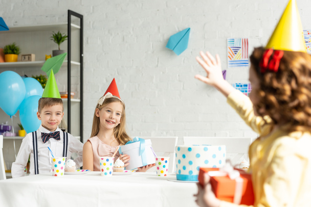 kid waving to friends in party caps sitting at table during birthday celebration at home - Фото, изображение