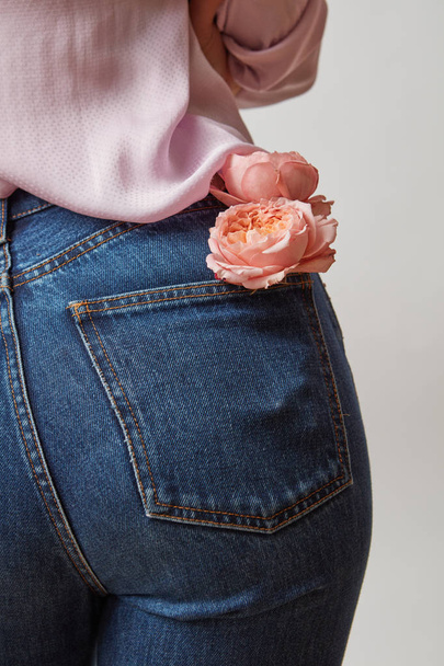 Beautiful female butt in jeans with fresh roses of coral living color in a back pocket on a light gray background. Place for text. - Foto, imagen