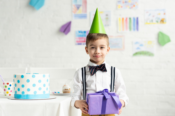 adorable boy in party cap looking at camera and holding present during birthday celebration - Valokuva, kuva