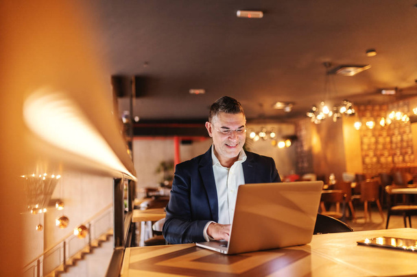 Sonriente hombre de mediana edad vestido casual inteligente usando el ordenador portátil y sentado en la cafetería
. - Foto, imagen
