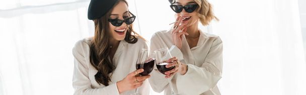 tiro panorâmico de morena feliz elegante e mulheres loiras em boinas pretas e óculos de sol rindo, fumando e bebendo vinho tinto
 - Foto, Imagem
