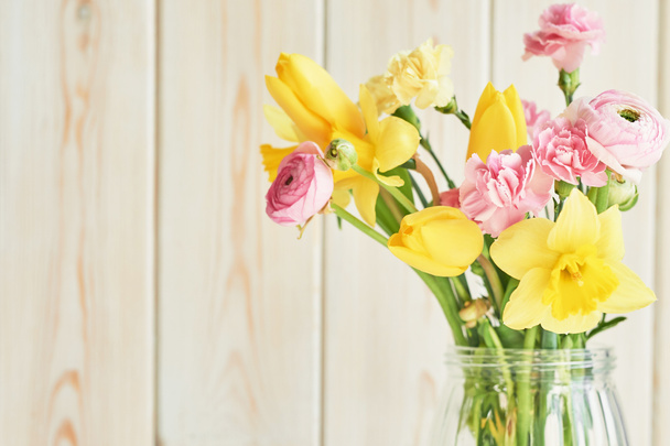 bouquet of spring flowers: tulips, carnations, ranunculi and daffodils in a vase on a table on a white wooden background - Photo, Image