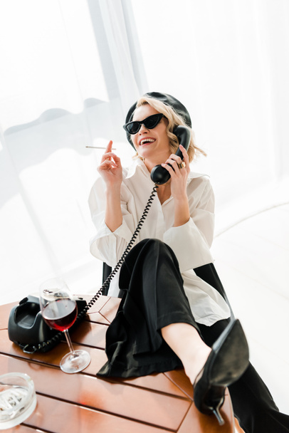 elegant blonde woman in black beret and sunglasses sitting with leg on table, smoking, laughing and talking retro phone - Photo, Image