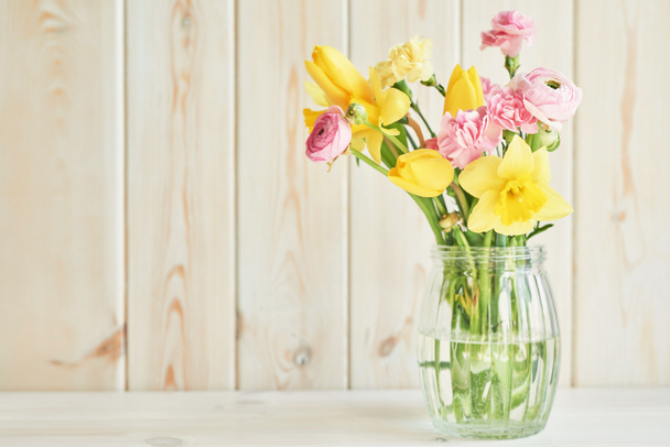 kukkakimppu: tulppaanit, neilikat, ranunculi ja narsissit maljakko pöydällä valkoisella puupohjalla
 - Valokuva, kuva