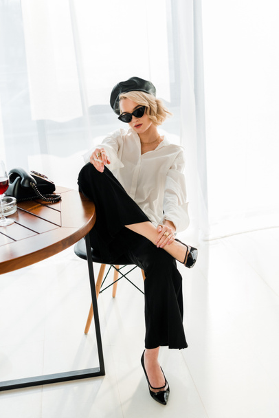 elegant blonde woman in black beret and sunglasses sitting on chair in pose and smoking cigarette - 写真・画像