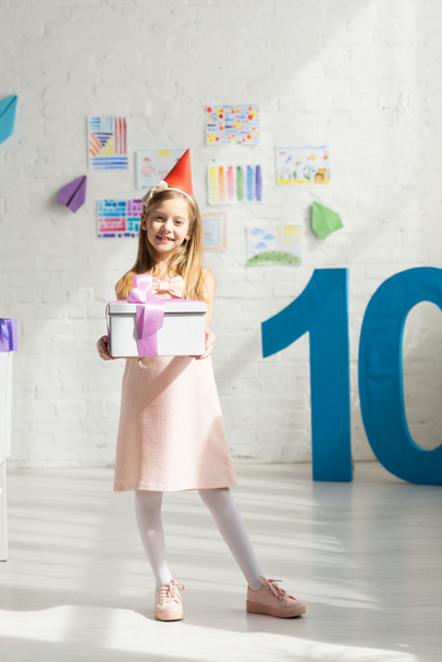 adorable happy kid in party cap holding present and posing near decorative number 10 - Фото, зображення