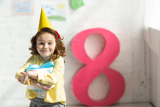 Очаровательный счастливый ребенок в кепке с подарком и позирует рядом с декоративным розовым номером 8
 - Фото, изображение