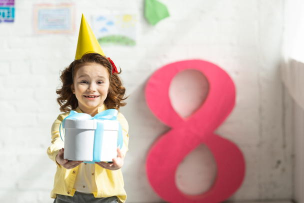 adorable smiling kid in party cap holding present and posing near decorative pink number 8 - Foto, Imagen