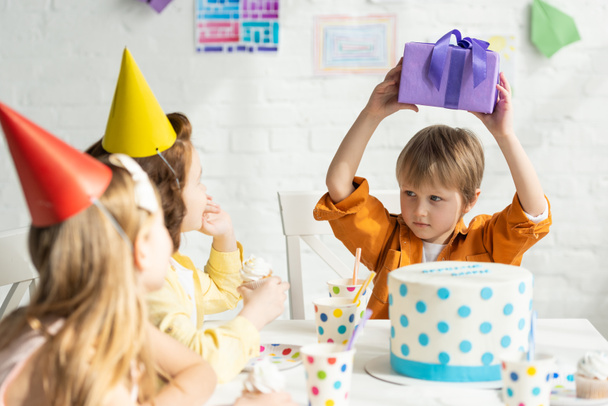 хлопчик тримає подарунок, сидячи за столом з друзями під час святкування дня народження
 - Фото, зображення