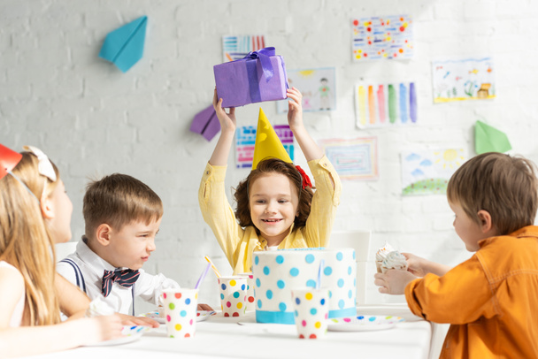 happy kid holding present while sitting at table with friends during birthday party celebration - Foto, Bild