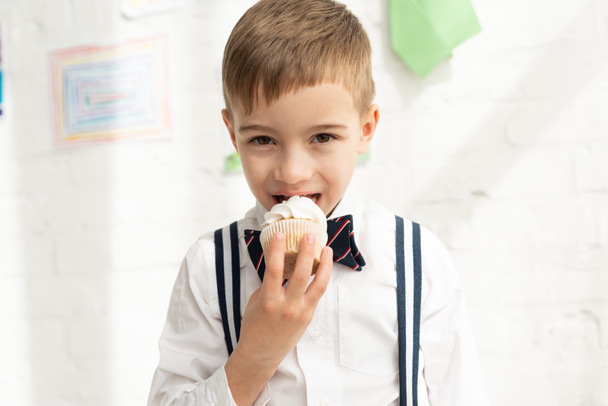 adorable preteen boy in bow tie eating cupcake and looking at camera - Photo, Image