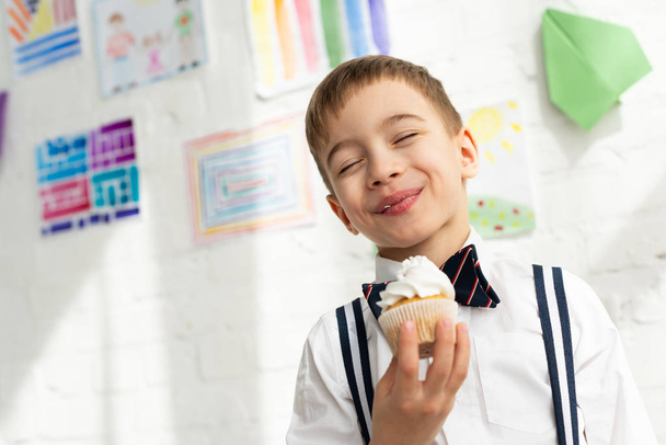 adorable preteen boy in bow tie holding cupcake and smiling with eyes closed - Photo, Image