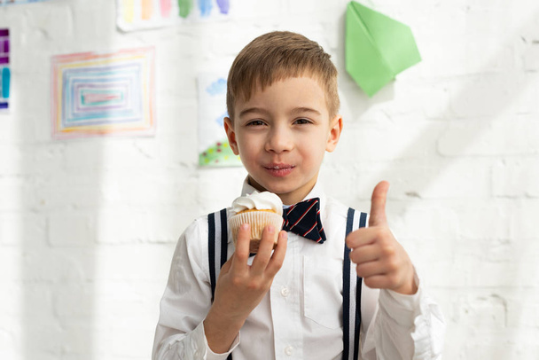 imádnivaló preteen fiú csokornyakkendő gazdaság Cupcake és mutatja hüvelykujjával fel jel - Fotó, kép
